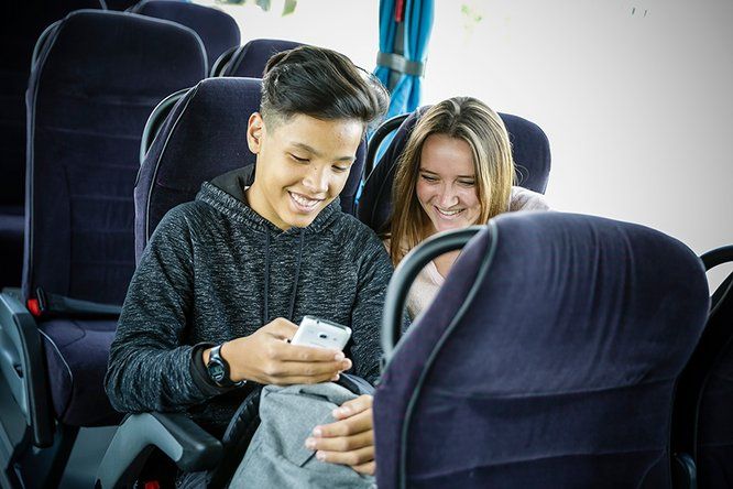 2 lycéens assis dans le car regardent un téléphone et sourient 